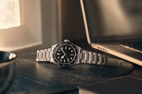 A stainless steel luxury dive watch on a slate surface beside a softly lit open laptop